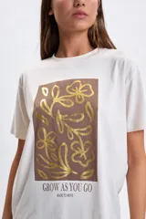 Remera de manga corta color crudo con estampa frontal rectangular en tonos marrones y dorados con diseño floral y la frase 'Grow as you go'.