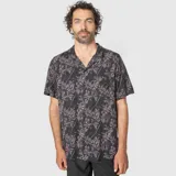 Camisa de manga corta con cuello cubano, confeccionada en viscosa. Presenta un estampado botánico con hojas en tonos oscuros, violetas y líneas blancas sobre fondo negro. Incluye un bolsillo frontal en el pecho.