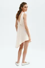 Vestido midi color beige de viscosa, sin mangas, con escote en V profundo adelante y atrás decorado con ribetes de encaje. Presenta una falda acampanada con bajo asimétrico y costuras en zigzag.