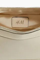 Cartera de hombro color crema, con diseño estructurado, cierre superior con tirador largo y correa ajustable con hebillas metálicas laterales.