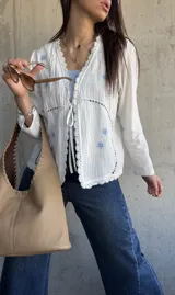 Capa blanca de manga larga con detalles bordados florales celestes y terminaciones de encaje en los bordes y la abertura frontal con tiras para atar.