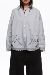 Camisa de manga larga color celeste, de corte holgado, con cuello clásico, cierre frontal de botones, bolsillo en el pecho y detalles de bordados florales y encaje en el ruedo y los puños.