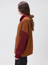 Pullover de tejido acanalado grueso con cuello alto y cierre frontal parcial. Presenta un diseño de bloques de color, con cuerpo en tono beige y mangas en contraste con rayas verticales en tonos rojizos. Incluye un detalle bordado en el pecho.