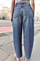 Jeans de tiro alto con diseño barrel, confeccionados en denim azul. Presentan cinco bolsillos, cierre frontal con botones y detalle de costuras verticales en el frente y la espalda. Su corte es holgado en la pernera y se estrecha hacia el tobillo.