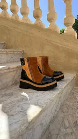 Botas cortas de cuero combinado en tonos tostado, marrón y detalles en charol negro. Cuentan con suela chunky, taco de 6 cm, cierre lateral y laterales de gamuza.