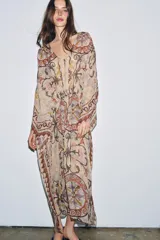 Vestido tipo túnica de viscosa con escote en pico y mangas tipo capa. Presenta un diseño estampado con motivos ornamentales en tonos tierra, beige y rosado, con pliegues en la cintura y bajo asimétrico.