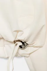 Chaqueta corta de nylon color beige, con cuello subido, capucha y cierre frontal con cremallera. Presenta mangas largas con puños elásticos, bolsillos laterales y detalles de cordones ajustables con lazadas en el pecho y el bajo.