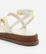 Sandalia plana tipo salomé color beige, con tiras finas, cierre con hebilla lateral y detalle decorativo dorado en la parte frontal. Cuenta con una suela de plataforma baja en tono madera.