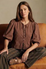 Blusa de algodón color marrón con diseño de bordado inglés (broderie) floral. Presenta cuello redondo con lazo de ajuste, cierre frontal con botones y mangas tres cuartos abullonadas con puños elásticos.