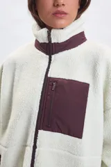 Campera de sherpa color crudo con cuello alto, cierre frontal completo y detalles en color bordó. Presenta un bolsillo frontal con cierre y dos bolsillos laterales, todos con terminaciones en contraste.
