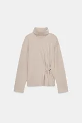 Top de cuello alto y manga larga, confeccionado en tejido de algodón. Presenta un diseño con frunces laterales y un aplique metálico decorativo.