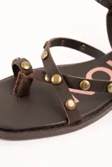 Sandalias planas de cuero marrón, con tiras finas decoradas con tachas metálicas redondas y tira ajustable alrededor del tobillo.
