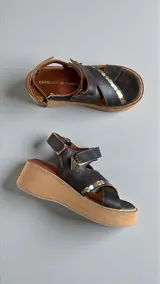 Sandalias de cuero color marrón oscuro con plataforma y taco de 5 cm. Tienen tiras cruzadas en el empeine con apliques metálicos decorativos y un dije colgante en forma de triángulo, además de una tira con cierre ajustable en el talón.