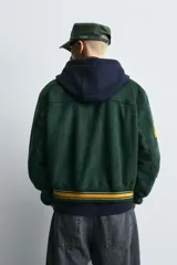 Cazadora tipo varsity de corte relajado confeccionada en tejido efecto ante color verde oscuro. Presenta cuello solapa, cierre frontal con botones a presión y puños y bajo acanalados con rayas en contraste. Incluye parches bordados en color amarillo en el frente y mangas.