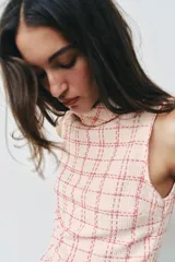 Vestido corto sin mangas de tejido texturizado con diseño de cuadros en tonos crudo y fucsia. Presenta cuello subido, trabillas decorativas con botones dorados en los hombros y cierre lateral con cremallera oculta.