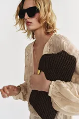 Bolso tipo sobre color marrón, con cuerpo trenzado y asa de mano rígida dorada. Incluye correa bandolera de cadena extraíble y cierre magnético.