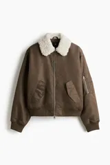 Campera bomber de nylon color marrón, ligeramente acolchada, con cuello de peluche blanco y cierre frontal. Incluye un bolsillo con cierre en una manga, bolsillos laterales con solapa y botón a presión oculto, y un bolsillo interior con botón a presión. Tiene remate acanalado ancho en puños y bajo, y un corte holgado.