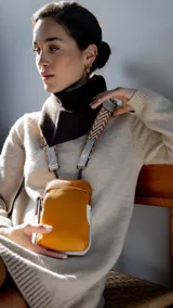 Phone bag color mostaza, confeccionada en símil cuero, con bolsillo frontal y correa de galón regulable y desmontable con diseño geométrico. Cuenta con cierre principal y espacio interior para celular y billetera.