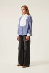 Camisa de popelina a rayas azules y blancas, de corte recto, con cuello y puños blancos. Presenta un babero plisado blanco.