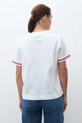 Remera blanca de algodón con cuello redondo y mangas cortas. Presenta bordados florales rojos en los hombros y en los bordes de las mangas.