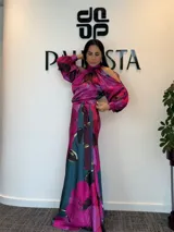 Vestido largo de satén con estampado floral en tonos fucsia, azul petróleo y negro. Presenta cuello alto, diseño con hombros descubiertos y mangas largas con puños. Incluye un lazo ajustable en la cintura para entallar la silueta.