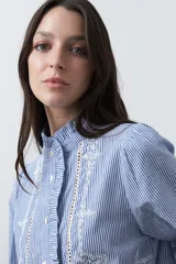 Camisa de rayas verticales con cuello mao, detalle de volados en el escote, bordados florales y recortes con calados. Posee mangas largas abullonadas con puños ajustados y cierre frontal con botones.