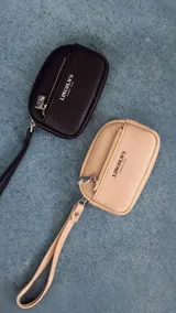 Monedero de mano con diseño compacto, confeccionado en símil cuero con textura granulada. Cuenta con un compartimento principal con cierre, un bolsillo frontal adicional y una correa de muñeca extraíble. Incluye el logo de la marca estampado en el frente.