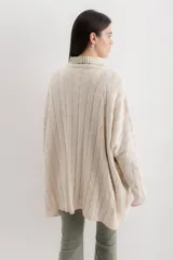 Sweater de punto color camel con diseño de trenzas verticales y acabado brillante. Presenta una silueta holgada tipo poncho, cuello redondo, mangas largas con puños ajustados y bolsillos frontales con detalle de etiqueta de cuero.