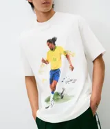 Remera de algodón con corte relaxed, cuello redondo y mangas cortas. Presenta un estampado frontal con una ilustración estilo acuarela de Ronaldinho jugando al fútbol.