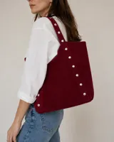 Cartera tipo tote bag color bordó, confeccionada en símil gamuza, con diseño amplio y detalle de tachas plateadas metálicas a lo largo de las tiras y los laterales.