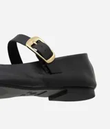 Zapatilla tipo bailarina de cuero negro, con punta redondeada, tacón bajo y una tira decorativa en el empeine con hebilla metálica dorada.
