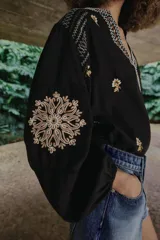 Camisa negra de manga larga con cuello redondo y escote en V, adornada con bordados florales en contraste en tonos beige y marrón a lo largo de la abertura frontal, hombros y mangas.