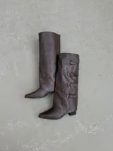 Bota de caña alta estilo slouchy color marrón, confeccionada en material sintético con acabado mate. Presenta un diseño de caña arrugada con cuatro correas horizontales ajustables mediante hebillas metálicas doradas y punta redondeada.