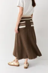 Falda midi de lana en color marrón, con diseño acampanado y tejido acanalado. Presenta una cintura elástica con cordón de ajuste, detalles de rayas en tonos beige y blanco en la parte superior, y costuras decorativas en el ruedo.