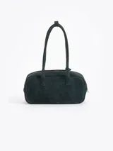 Cartera de mano de estructura rectangular, confeccionada en cuero descarne vacuno color verde oscuro. Presenta doble manija larga, cierre superior y un charm metálico dorado en forma de vaquita de San Antonio colgando de un tag lateral. Incluye el logo de la marca grabado en relieve en el frente.