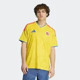 Camiseta de fútbol Adidas de la selección de Colombia, modelo Primera Equipación 26, color amarillo con un patrón sutil en relieve. Presenta cuello en V con ribete azul, logo de Adidas en azul y escudo de la federación colombiana. Las tres franjas de Adidas en los hombros son rojas y azules.