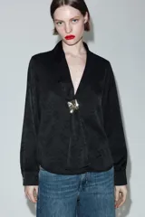 Camisa de satén negro con diseño jacquard de flores tono sobre tono. Presenta escote en pico con solapas, cierre frontal con botones y un broche metálico decorativo en el centro. Tiene mangas largas con puños abotonados.
