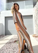 Vestido largo de tejido de punto con diseño geométrico en tonos tierra, negro y blanco. Presenta escote en V profundo, mangas tres cuartos y tajos laterales en el ruedo.