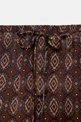 Pantalón de tiro alto confeccionado en seda, con cintura ajustable mediante cordones. Presenta un diseño de patchwork con diversos estampados geométricos y paisley en tonos tierra, verde y violeta.