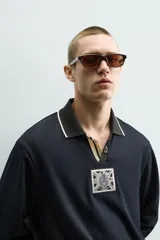 Polo de manga larga en tejido técnico acanalado color negro, con cuello solapa y abertura frontal. Presenta puños con ribete a contraste y una etiqueta aplicada en el pecho con el diseño conmemorativo de la Copa Mundial de la FIFA 1966.