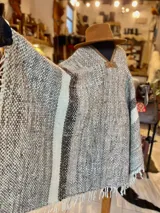 Poncho de lana virgen tejido en telar con diseño geométrico a rayas en tonos gris, marrón y celeste, con apliques de cuero en el escote y flecos en el ruedo.