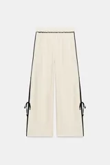 Pantalón ancho de tiro medio confeccionado en lino, color crudo con detalles de vivos negros a contraste en los laterales y la cintura, con aberturas y lazos en los bajos.