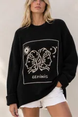 Sweater de punto negro con diseño oversize, cuello redondo y manga larga. Presenta un bordado frontal en color crudo con motivo del signo del zodíaco Géminis, acompañado de detalles de sol, luna y estrellas dentro de un recuadro.