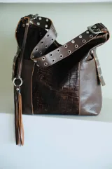 Cartera tipo bolso de cuero marrón con textura croco combinada con cuero liso, con correa ajustable decorada con ojalillos metálicos y un detalle de flecos colgantes con argolla plateada.