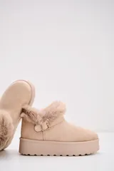 Bota corta tipo moon boot color beige, confeccionada en gamuza sintética con interior y borde superior de piel sintética. Presenta una suela de plataforma plana y una tira decorativa con hebilla metálica en el lateral.