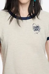Remera corta de corte ajustado, color beige claro con finas rayas horizontales oscuras. Posee cuello redondo y ribetes en contraste de color azul marino en el cuello y los bordes de las mangas cortas. Lleva un pequeño bordado en forma de corazón con texto en el pecho.