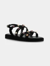 Sandalias planas de cuero gamuza en color negro, con múltiples tiras cruzadas decoradas con tachas metálicas doradas y aplicaciones de piedras rojizas. Cuenta con suela de PVC y tira ajustable en el talón con hebilla metálica dorada.