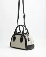 Cartera tipo baúl bicolor, combinando cuerpo de lona beige con detalles de símil cuero negro en las asas, bordes y correa. Cuenta con doble asa de mano, cierre superior y correa ajustable y desmontable para usar al hombro o cruzada.