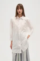 Camisa blanca de manga larga, corte holgado y largo, con cuello camisero y botones frontales. Presenta bordados florales en tono blanco sobre el tejido.