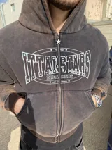 Campera con capucha y cierre frontal de color marrón con efecto acid wash y desgaste. Presenta un estampado gráfico frontal en color blanco con inscripciones y detalles de pedrería, además de bolsillos tipo canguro y un tirador de cierre en forma de estrella.